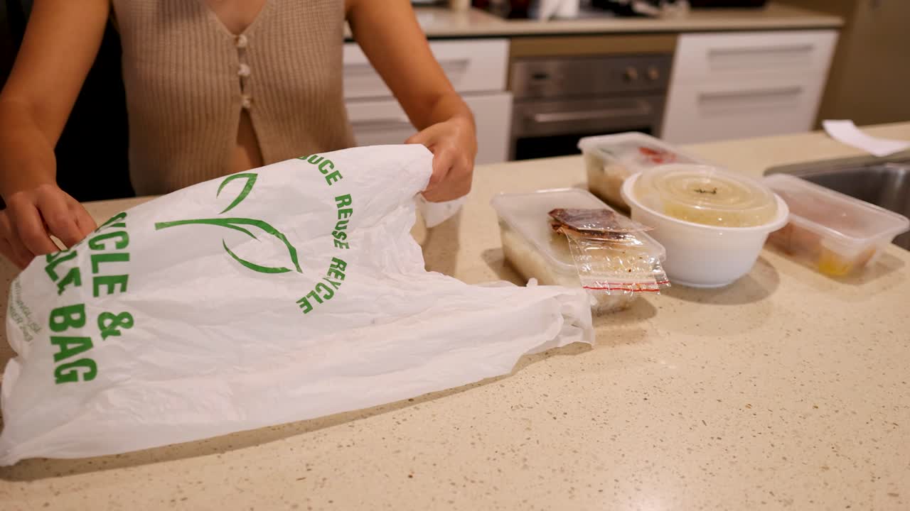 Hands organize food containers into a recyclable bag on a kitchen counter under warm lighting