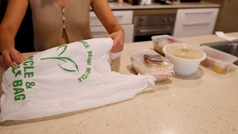 hands organize food containers into a recyclable bag on a kitchen counter under warm lighting