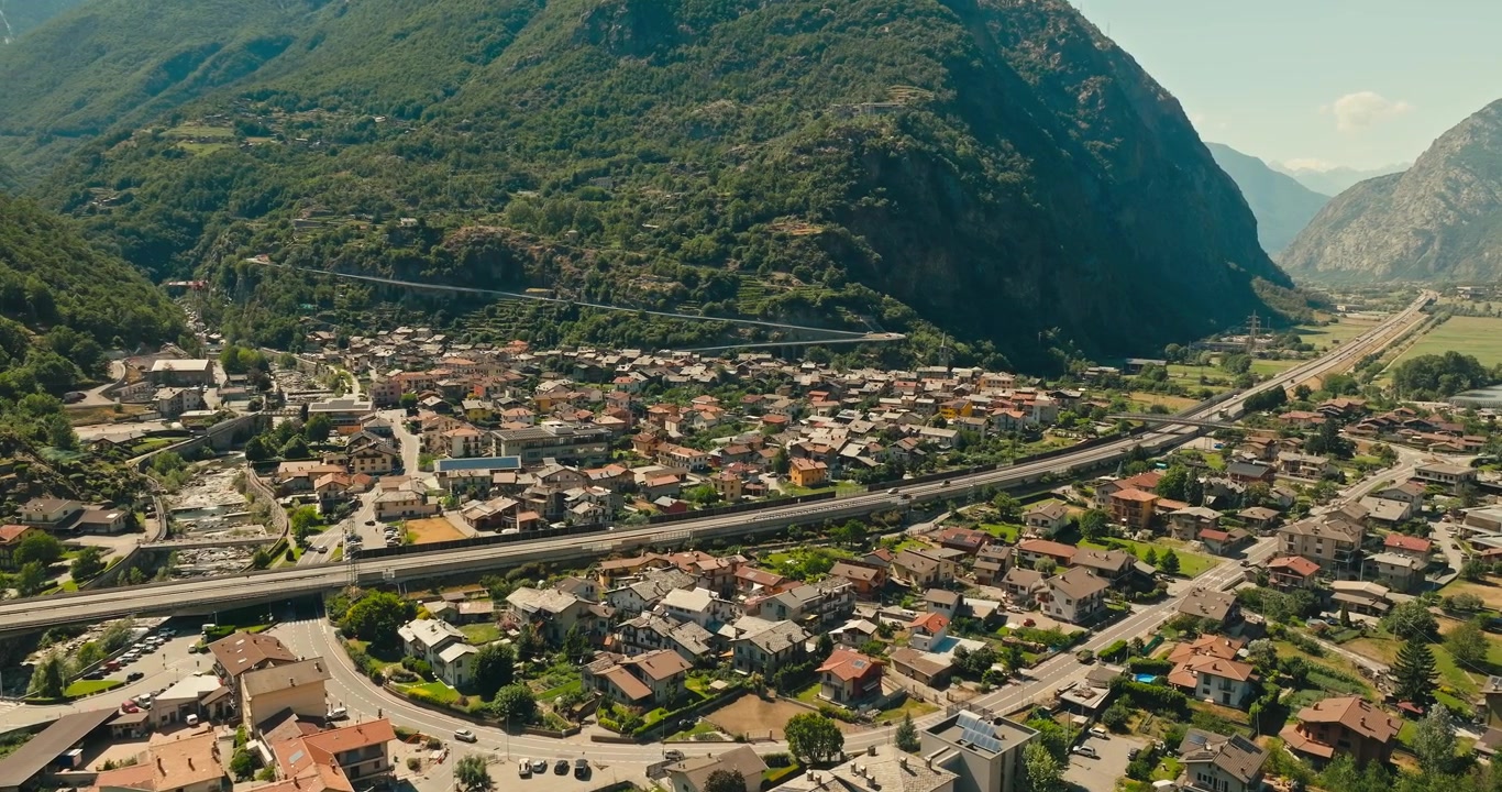 Aosta Vadisi'nde berrak mavi bir yaz gökyüzünün altında muhteşem dağlarla çevrili, otoyol boyunca seyahat eden arabaların olduğu büyüleyici bir İtalyan kasabasının drone görüntüsü. Manzaralı Avrupa altyapısı.