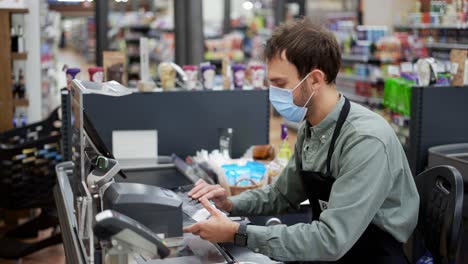 Sales-clerk-in-mask-working-at-grocery-store.-Male-cashier-in-black-apron-at-cash-desk-and-unrecognizable-customer
