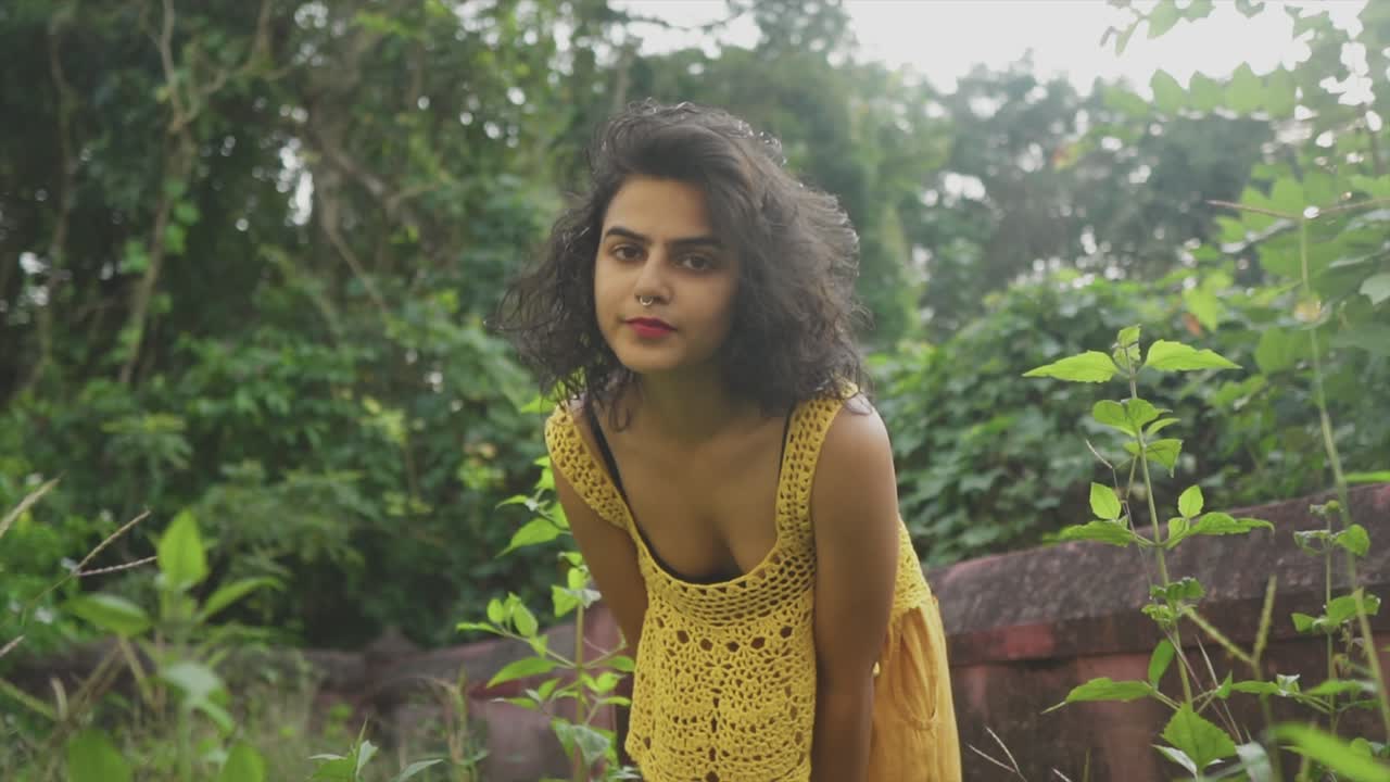 uma atraente mulher asiática vestindo um top de crochê amarelo curvando-se e jogando o cabelo para trás enquanto ela olha para cima ao ar livre em um jardim cercado por uma vegetação luxuriante