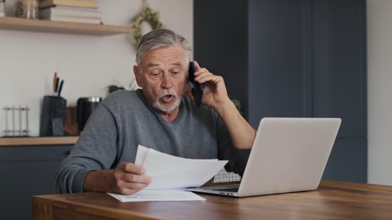 un anciano caucásico en shock mirando la computadora portátil y haciendo una llamada telefónica.
