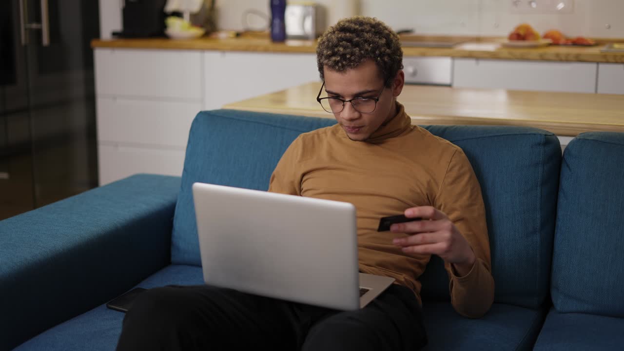 Young man buying online uses a laptop and a credit card on sofa home