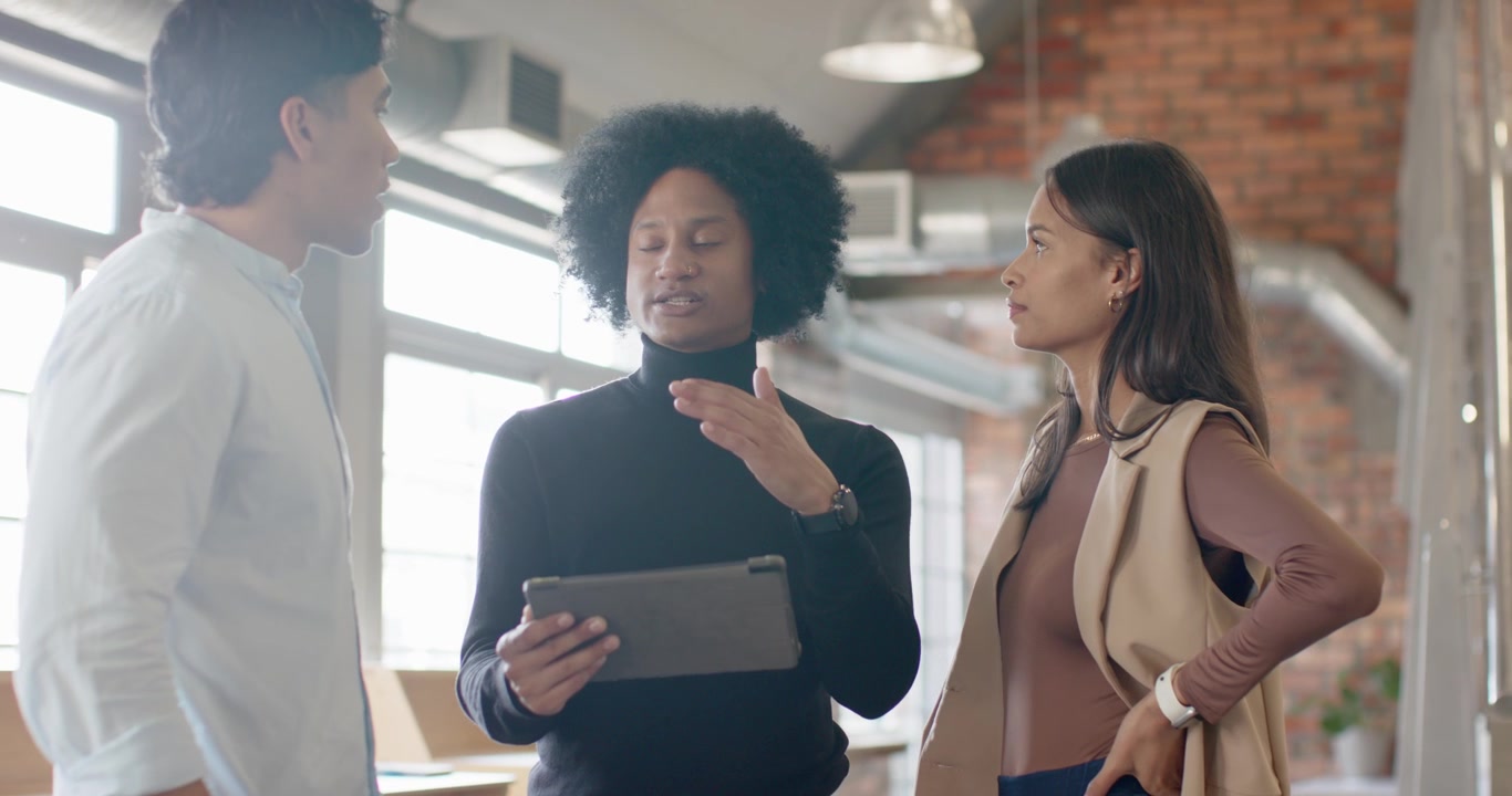 Diverse coworkers discussing as center colleague tapping tablet to review project near desks
