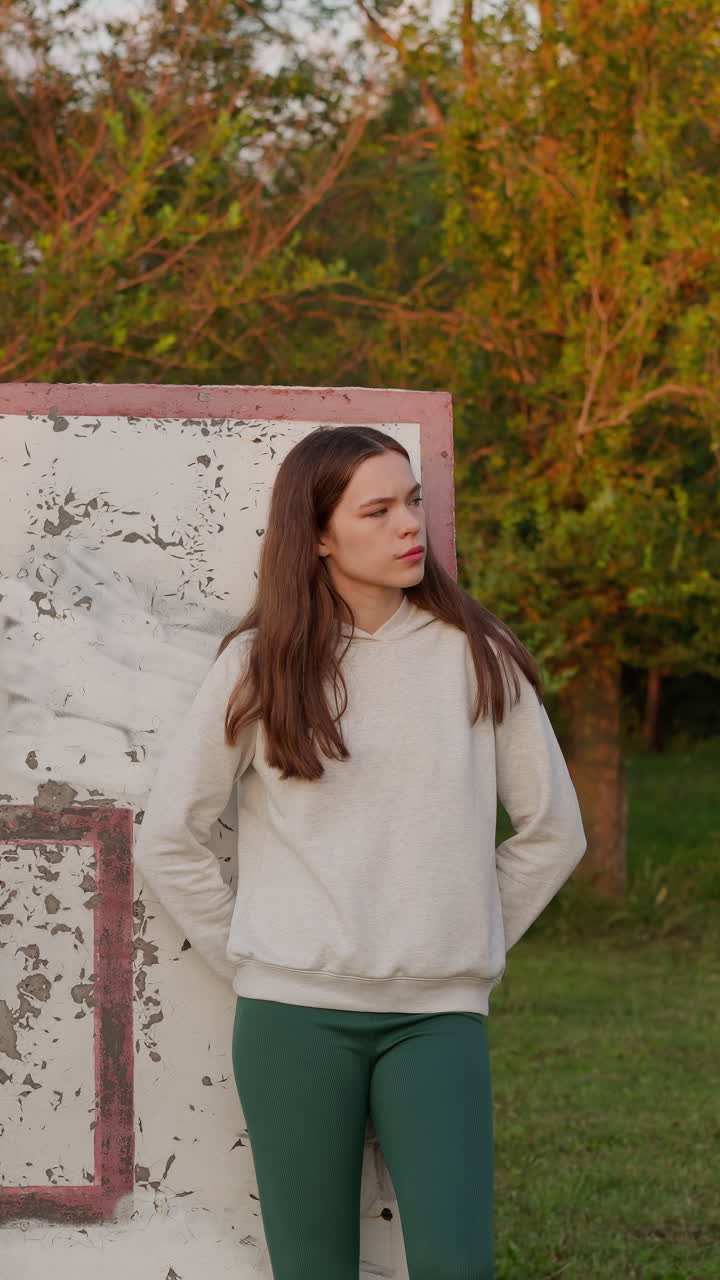 una mujer vestida con ropa deportiva está de pie en una cancha de baloncesto abandonada rodeada de hierba verde.
