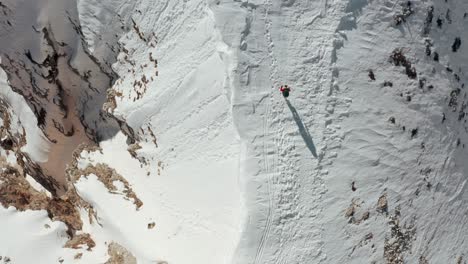 Toma-Aérea-De-Drones-De-Un-Excursionista-Caminando-A-Lo-Largo-De-Una-Cordillera-Nevada-Con-Vistas-A-Una-Caída-Empinada