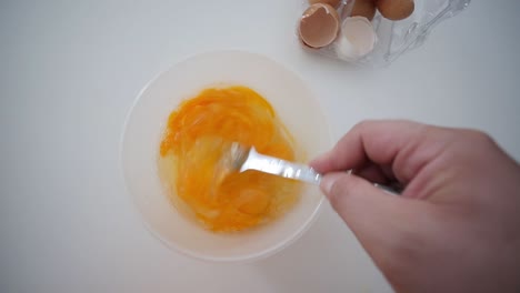 whisking egg yolks with fork in bowl top view with broken egg shells in background