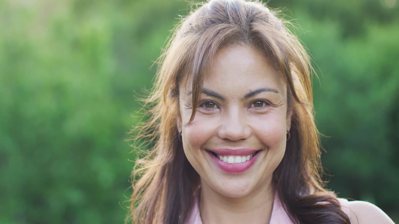 Smiling woman tilting right, blinking and keeping gaze in park, in light-pink blouse and studs