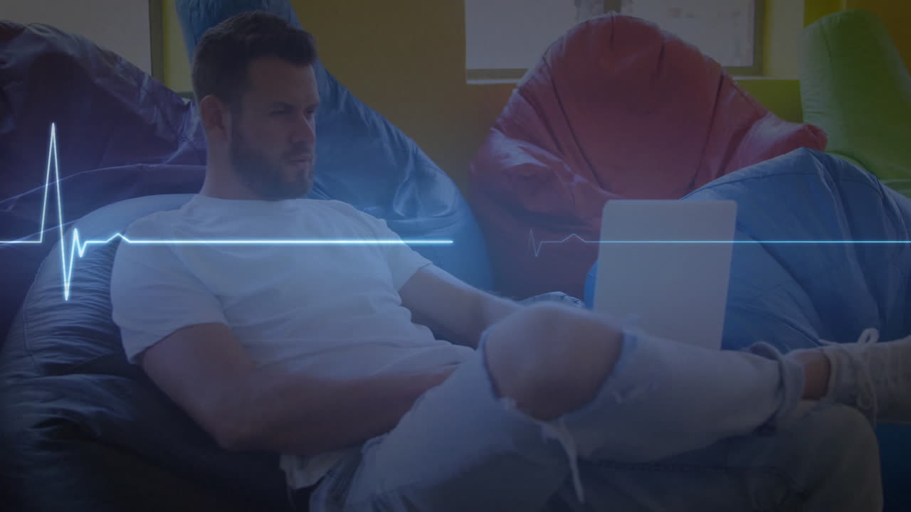 monitor de frecuencia cardíaca azul brillante contra hombre caucásico usando una computadora portátil sentado en una bolsa de frijoles en casa