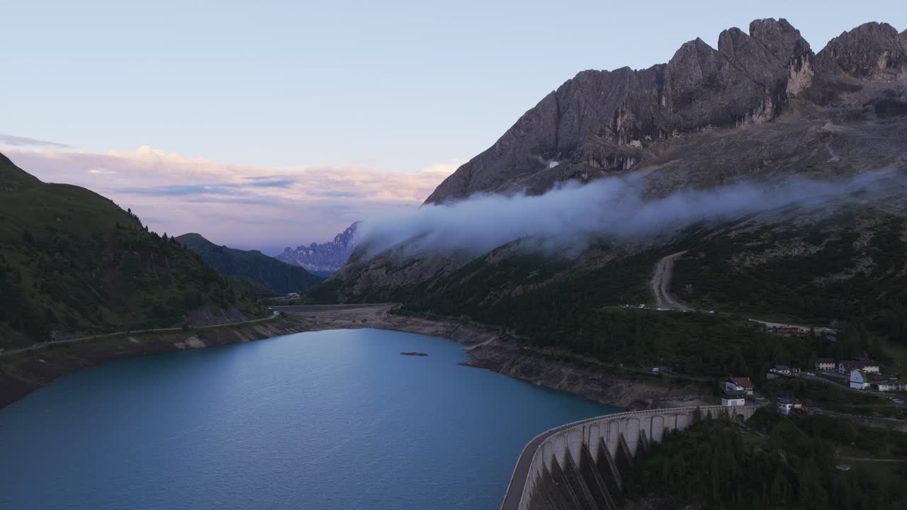 Fedaia Lake and dam at dusk, Marmolada mountain range with low clouds, Dolomites, Italy. Aerial drone panoramic view