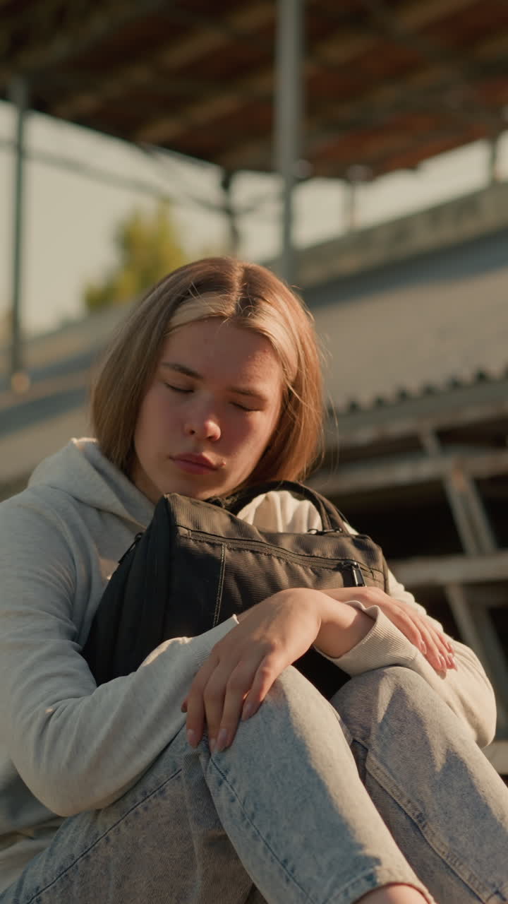Close-up of a young woman gazing thoughtfully while seated alone on empty stadium bleachers, hugging her bag tightly with a reflective expression, sunlight softly illuminates her face