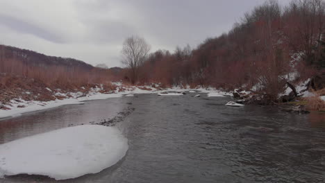 Drone-shot-over-a-almost-frozen-river-near-a-deciduous-forest