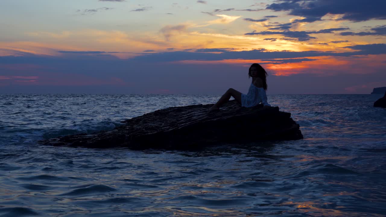 chica posando como una sirena sobre el gran acantilado bañado por el mar y mirando el hermoso cielo de la colorida puesta de sol