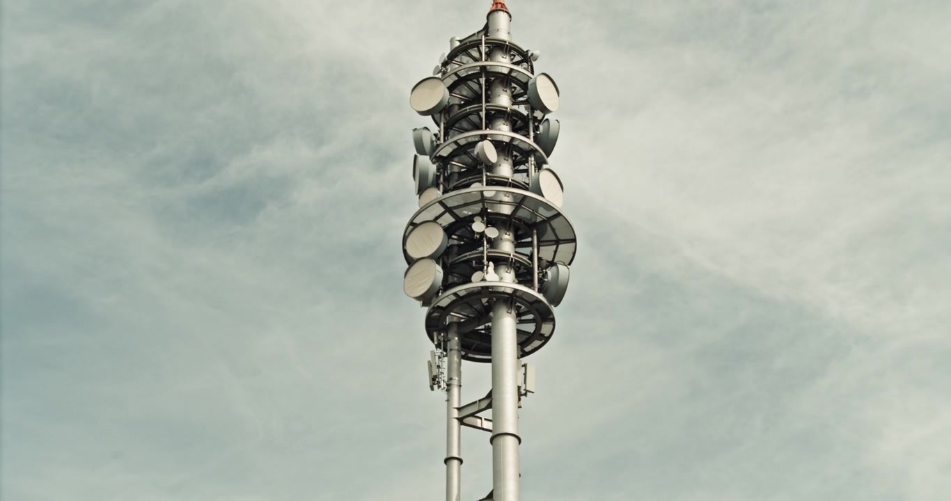 Zoom indietro da una vista ravvicinata delle numerose antenne e parabole a microonde su una torre di telecomunicazioni per rivelare l'intero traliccio contro un cielo grigio. Rete mobile e infrastruttura di comunicazione, Germania.