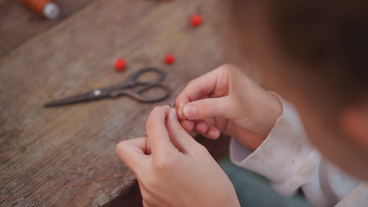 Closeup caucasian child threading beads. Playful outdoor craft moment on wooden bench with scattered red beads, scissors nearby, blurred face, nimble fingers assembling necklace, curiositydriven