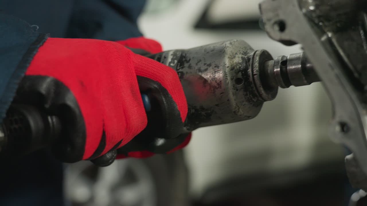 mecánico con guantes rojos opera una herramienta neumática en el bloque del motor en un taller industrial, un primer plano captura trabajos de reparación de precisión y componentes metálicos con un vehículo blanco borroso en el fondo