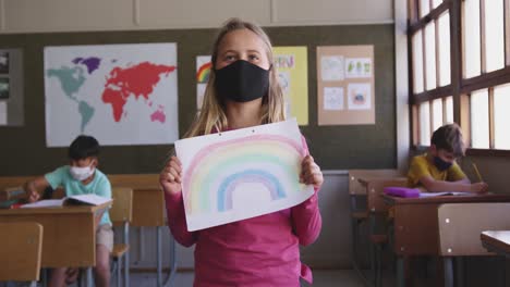 Girl-wearing-face-mask-holding-a-rainbow-painting-in-class-at-school-