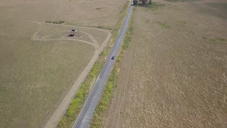 Toma-Aérea-De-4k-De-Un-Todoterreno-Plateado-Que-Conduce-Por-Una-Carretera-Rural-Larga-Y-Recta,-Aislada-Contra-Campos-Dorados-Y-Desolados-Mientras-La-Cámara-Retrocede-Y-Revela-Una-Amplia-Extensión-Y-Horizonte-Con-Colinas-Y-Cielo