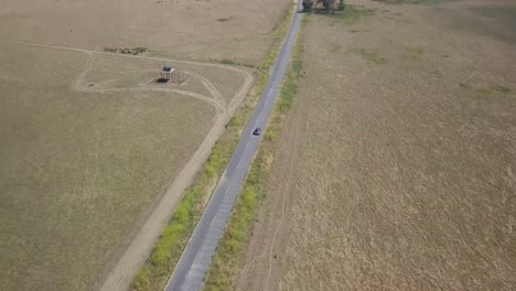 toma aérea de 4k de un todoterreno plateado que conduce por una carretera rural larga y recta, aislada contra campos dorados y desolados mientras la cámara retrocede y revela una amplia extensión y horizonte con colinas y cielo