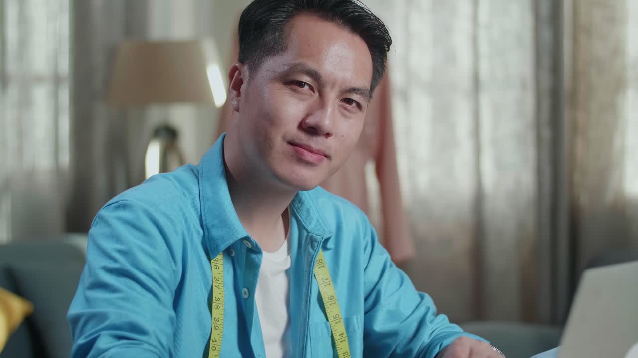 스튜디오에서 옷을 디자인하는 동안 무을 고 카메라에 미소 짓는 아시아 남성 디자이너의 클로즈업