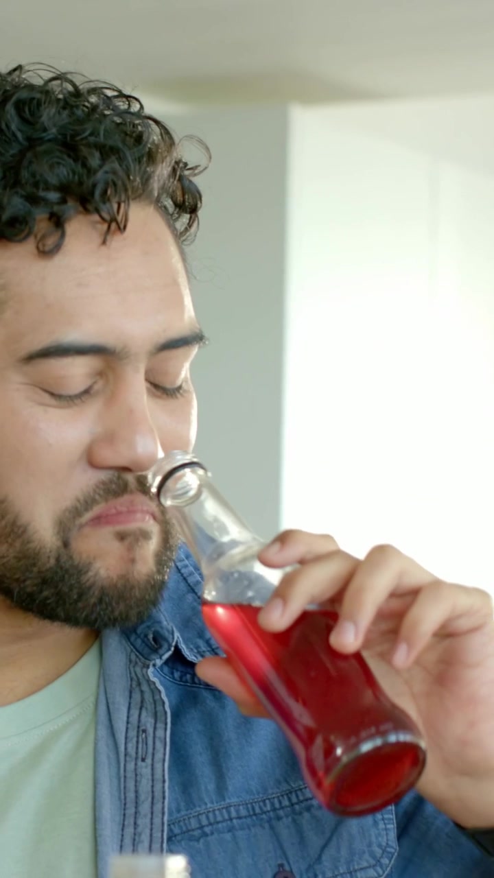 Vertical video: Inspecting bottle, denim-clad man sipping red drink tasting in kitchen, copy space