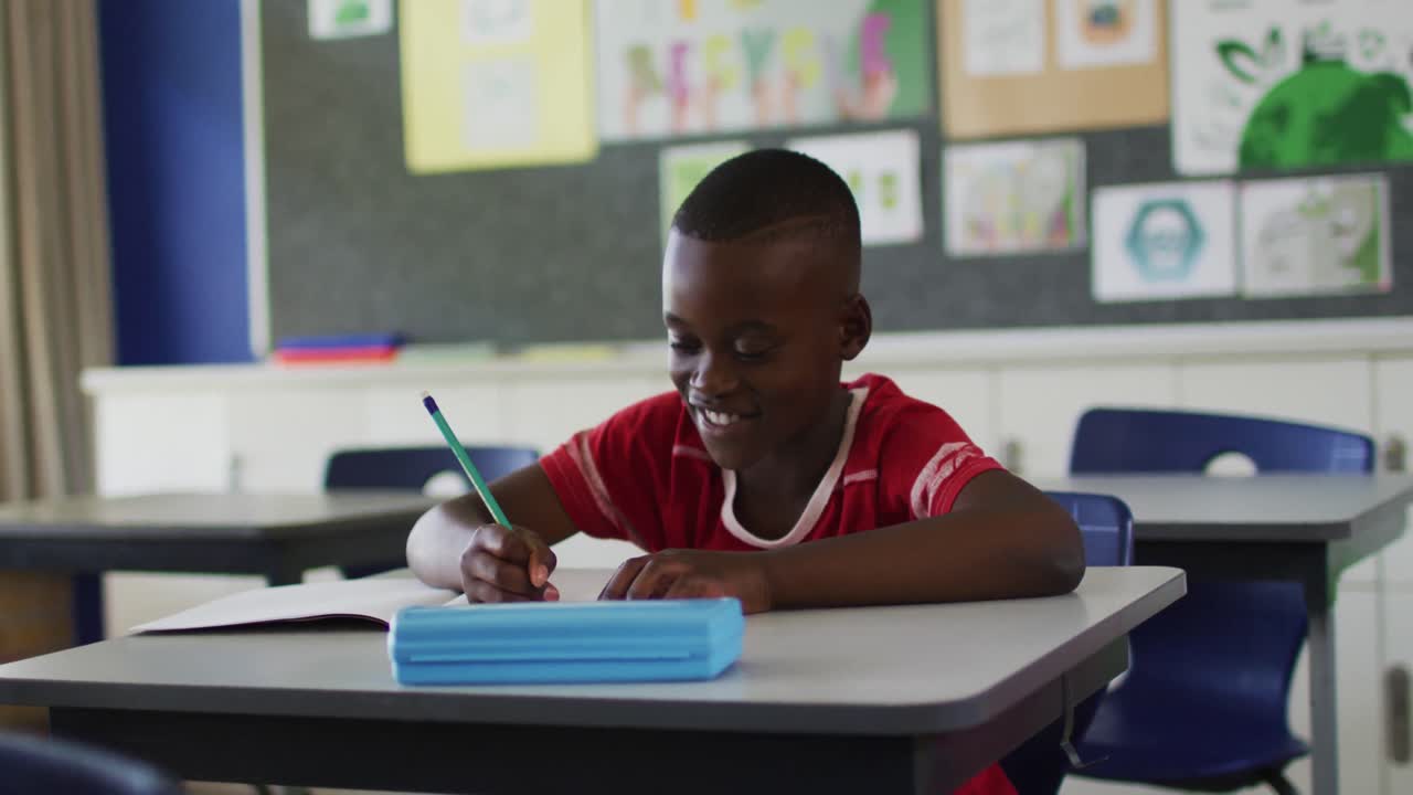Portrait of happy african american schoolboy sitting at classroom, making notes, looking at camera