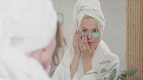 happy attractive woman in bath towel with cosmetic patches under eyes kooking at mirror and enjoying morning time at bright bathroom