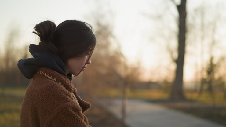 Close-up of a sorrowful girl walking through a park at sunset, wearing a brown coat and a hoodie. Her face is partially obscured by her hair, reflecting a quiet against the soft, fading light