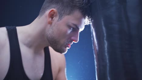 Tired-male-athlete-training-boxing-with-punching-bag-in-dark-gym.