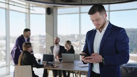 bearded caucasian man in official suit checking his smartphone. business team member standing at the foreground with phone in hands. colleagues working at the backdrop.