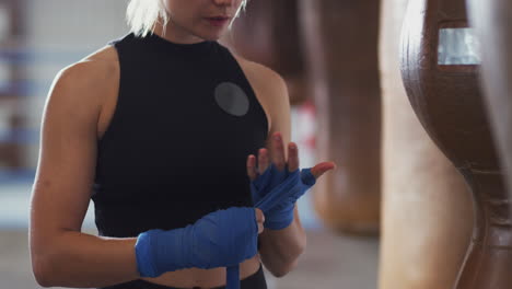 female boxer training in gym putting wraps on hands standing next to punching bag