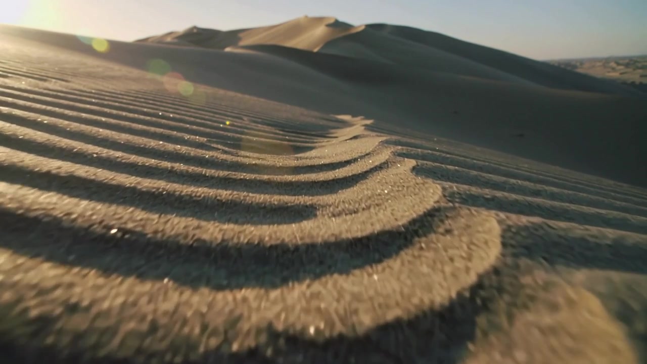 Con una ripresa dal basso, la telecamera mostra sabbia increspata che si muove attraverso il deserto verso le dune sotto un bagliore solare