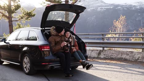 a loving couple in a road trip. the guy and the girl are sitting in the open trunk of the car, eating sandwiches. admiring the view around - beautiful nature of norway. stop outdoors in middle of the road