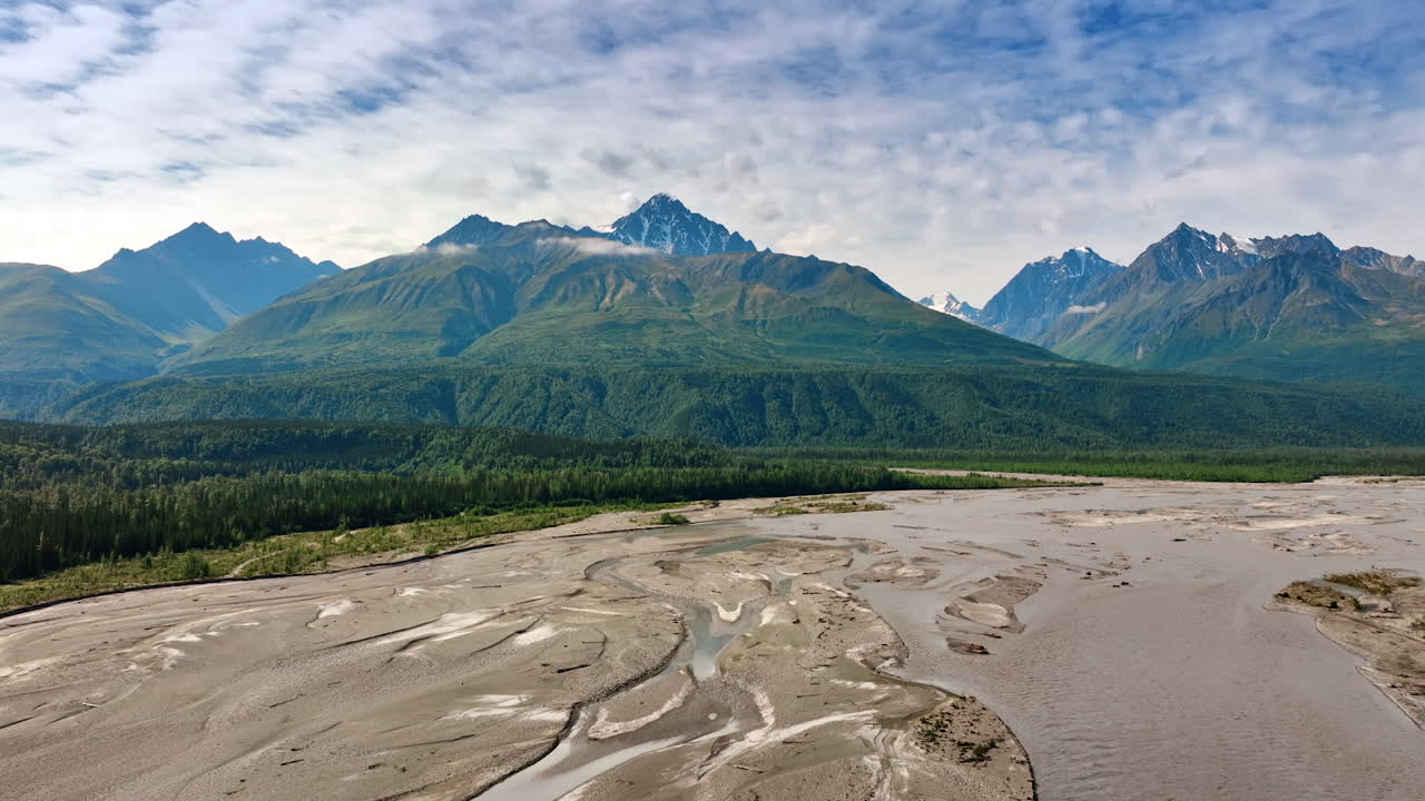 Going down over the delta of shallow river flowing in the wooded valley. Ridged verdant mountains at backdrop. Overcast sky above. Alaska, USA