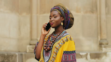 Beautiful-Young-Charming-Woman-In-The-Traditional-Outfit-And-With-Scarf-On-The-Head-Talking-Joyfully-On-The-Phone-On-The-Wall-Background