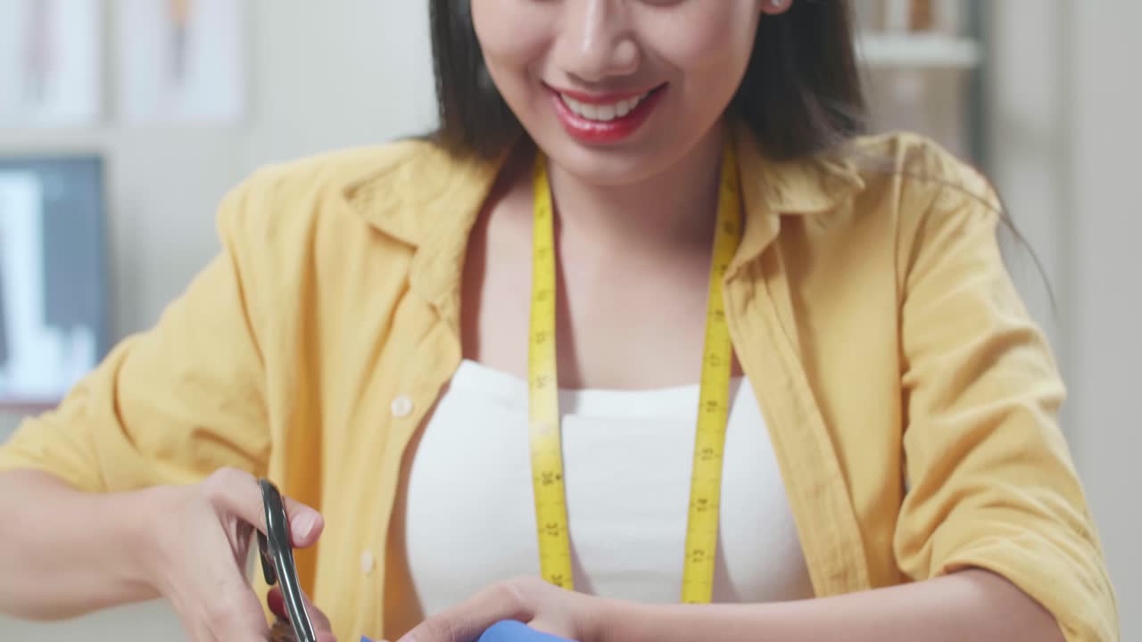 미소 짓는 아시아 여성 디자이너가 스튜디오에서 새로운 드레스를 만드는 동안 가위로 천을 자르고 있습니다.