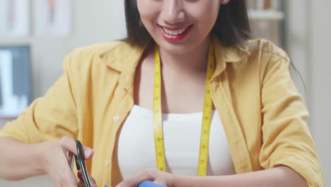 미소 짓는 아시아 여성 디자이너가 스튜디오에서 새로운 드레스를 만드는 동안 가위로 천을 자르고 있습니다.