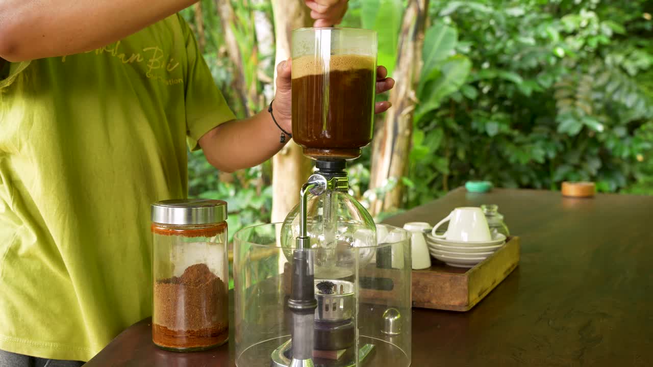 Barista brews coffee with a siphon extractor in a natural outdoor setting, showing the process of mixing, heating, and filtering grounds for a traditional handcrafted brew