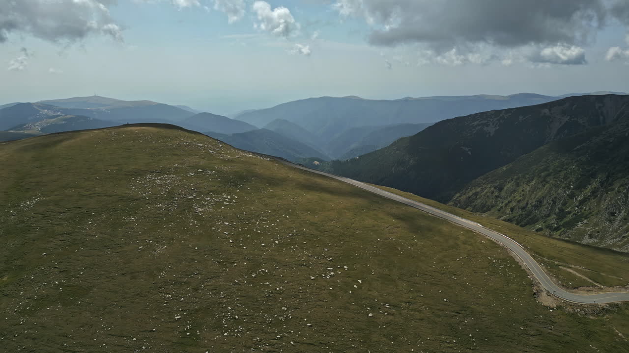 Majestic panorama of Romania's Transalpina, highlighting vast green plateaus and a solitary road carving its way through, with distant mountain layers under a dappled sky
