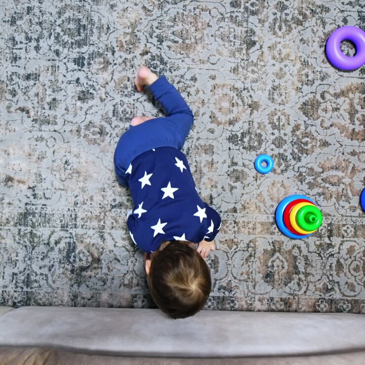 Tiny kid sitting on the floor puts the phone under sofa. Toy pyramid on the floor near the baby. View from top