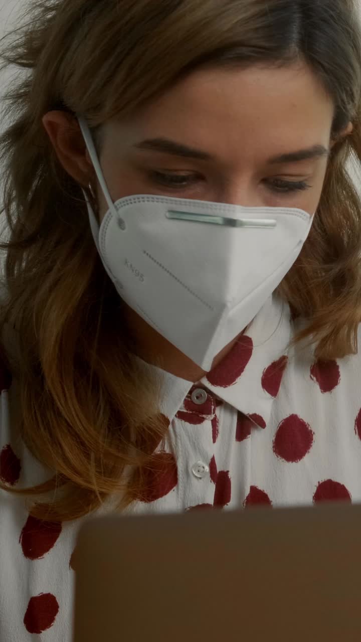 Giovane donna con mascherina protettiva che lavora al computer portatile in casa.