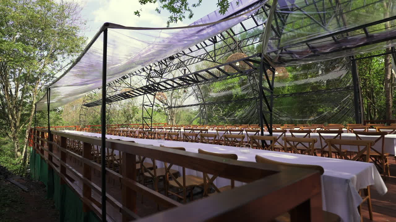 Slow motion reveal of a transparent gazebo wedding venue shows rows of white-covered tables and wooden chairs surrounded by lush greenery, blending formal elegance with natural forest ambiance