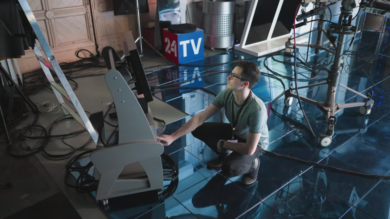 Man crouching and adjusting digital monitor and timer settings in modern studio, professional production environment with filming equipment, focused on workspace, tech action in progress
