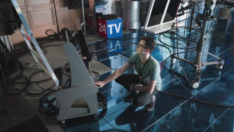 man crouching and adjusting digital monitor and timer settings in modern studio, professional production environment with filming equipment, focused on workspace, tech action in progress