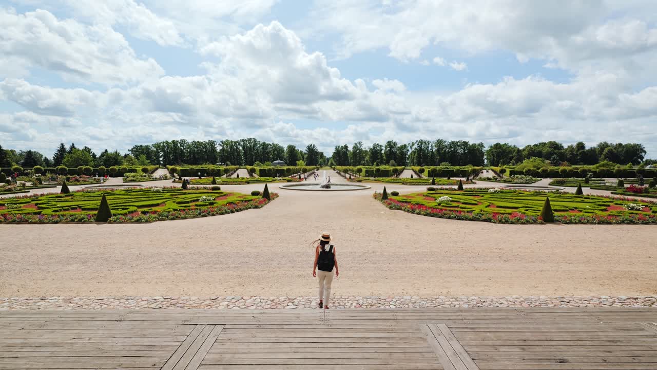 Elegant woman descends steps toward symmetrical French garden in summer breeze