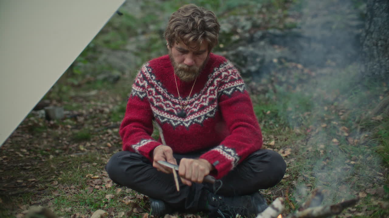 Man Whittling Wooden Stick while Sitting by Campfire under Tarp Shelter