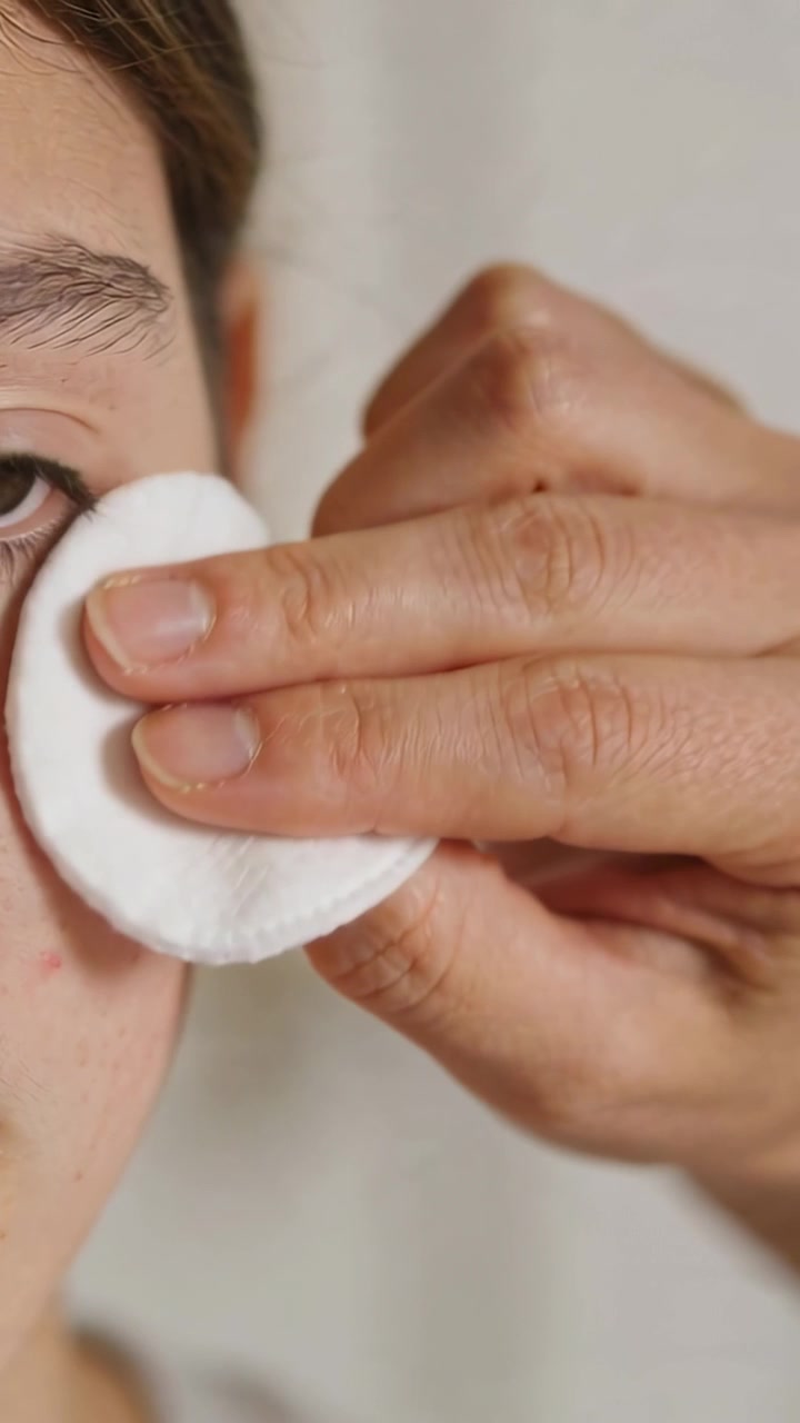 Vertical video: Pressing cotton pad, adult hand wiping teen's left cheek at home for skincare