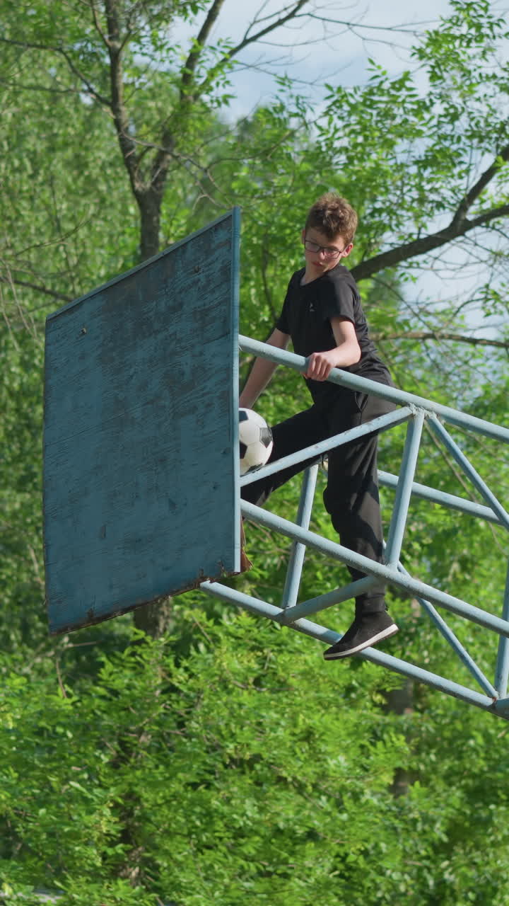 un niño está sentado en una estructura al aire libre de hierro azul, centrándose atentamente mientras se estira y mira hacia abajo, una pelota de fútbol desgastada está atascada en el marco de hierro