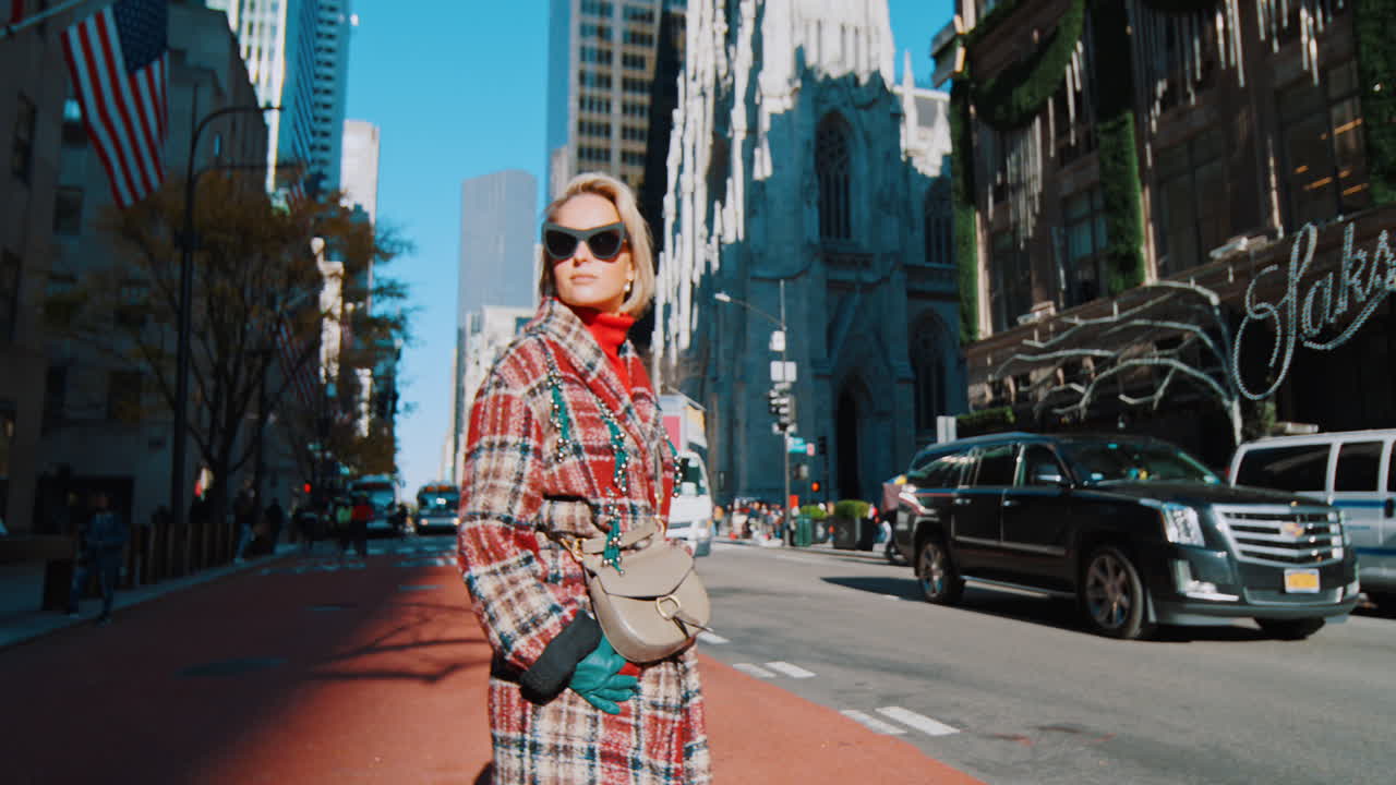New York, USA, 5 ottobre 2025: Donna con occhiali da sole in posa davanti a una cattedrale. Mezzo busto di donna con cappotto a quadri e occhiali da sole a occhi di gatto in piedi per una strada di New York