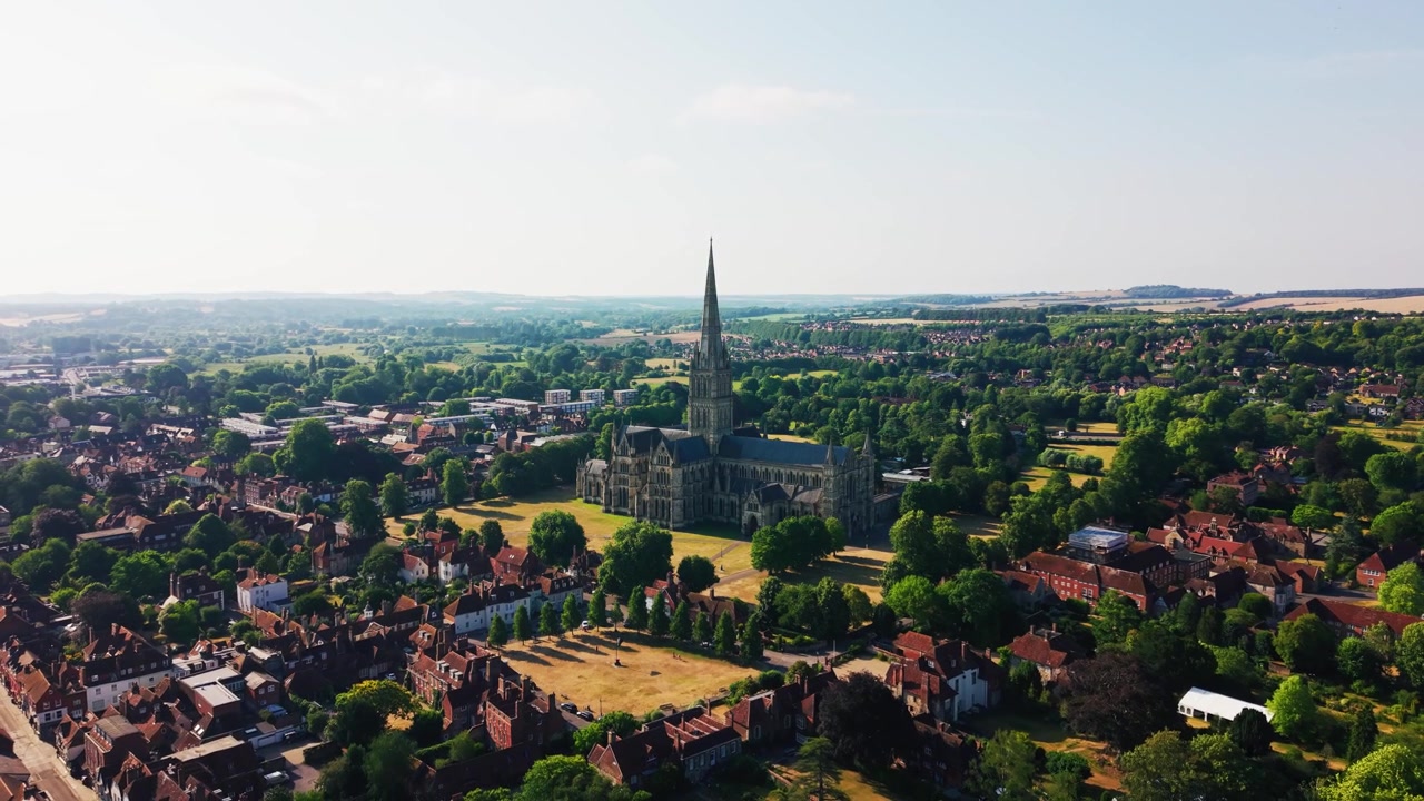 Vista dall'alto con drone della Cattedrale di Salisbury, che mostra la sua famosa guglia medievale, lo storico Cathedral Close, i prati verdi e il paesaggio urbano circostante.